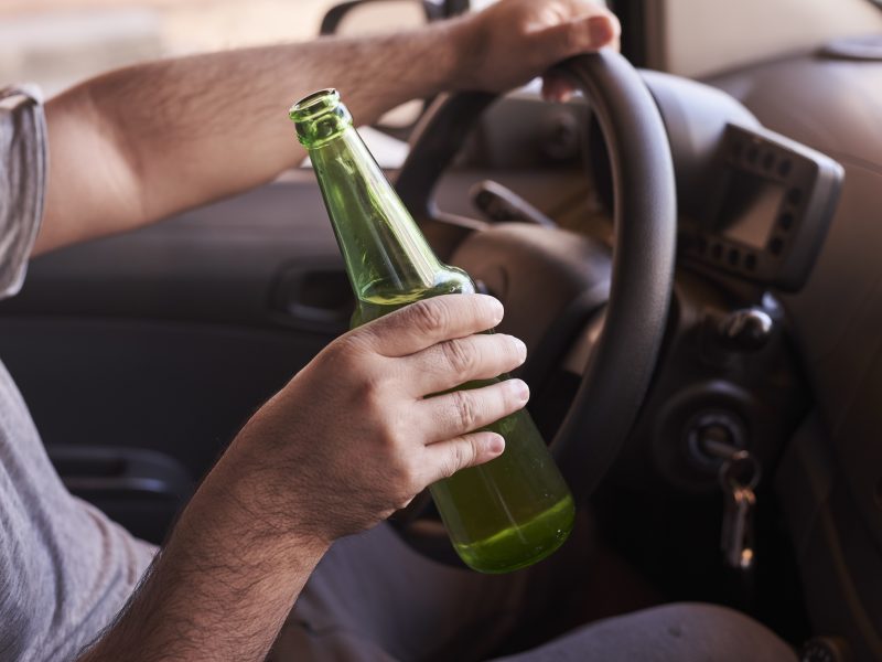 A bottle of beer in a man's hands driving the car during daytime